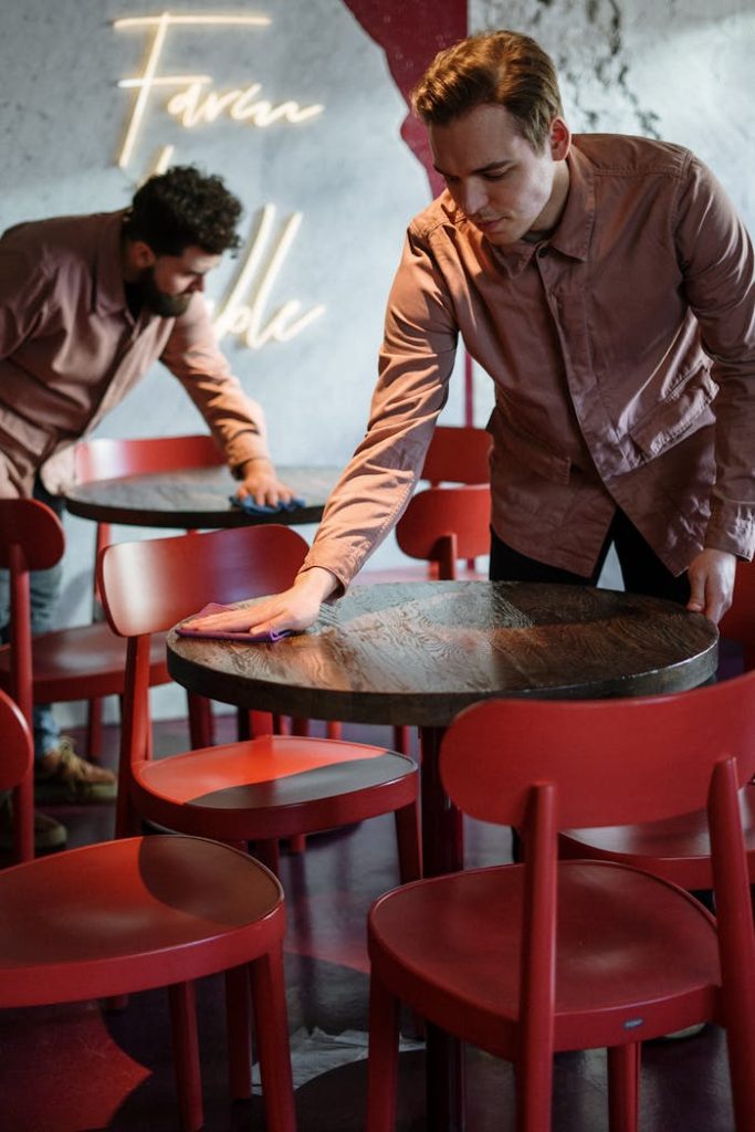 man-in-brown-leather-jacket-holding-brown-wooden-round-table-4254270 Two staff members prepare a modern cafe interior with red chairs and neon lighting.